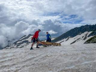 In den Alpen: Ein Mann und eine Frau mit einem Schlitten, auf dem sie Baumaterial über den Altschnee schieben.