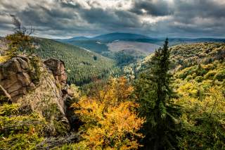 Landschaft mit Fels im Vordergrund und herbstlich gelb bewaldeten Bergen, darüber ein bedrohlicher Himmel.