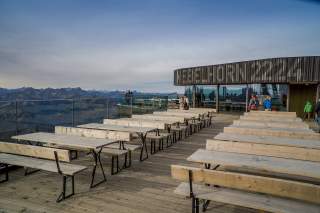 Eine Holzterrasse mit Holzbänken. Vor einem Gipfelrestaurant mit der Aufschrift 
