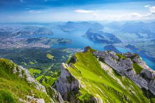 Blick von einem imposanten Gipfel mit steil abfallenden Hängen auf das Tal mit einem verzweigten See und anderen, niedrigeren Bergen.