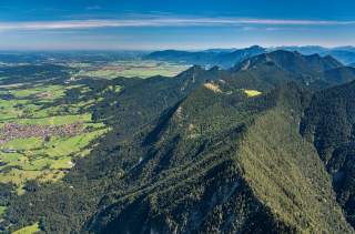 Luftaufnahme von bewaldeten Bergen mit grünen Tälern und einem Dorf am linken Bildrand unter blauem Himmel.