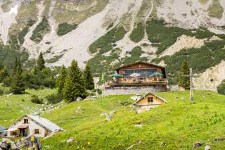 Berghütte mit Terrasse und mehreren Hütten auf einer grünen Almwiese vor steinigem Berghang, im Vordergrund ein Holzkreuz.