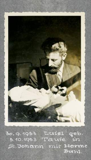 Schwarzweißfoto von einem Mann im Anzug mit Bart, der ein in eine Decke gewickeltes Baby auf dem Arm hält; darunter handschriftliche Notizen mit Daten und Namen.