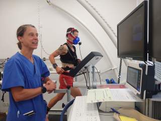 Eine Person läuft auf einem Laufband mit einer Atemmaske und Sensoren am Körper, während eine medizinische Fachkraft in blauer Kleidung die Untersuchung überwacht. Im Vordergrund sind Monitore und ein Computerarbeitsplatz zu sehen.