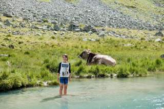 Ein Mädchen steht im klaren Wasser eines Gebirgsbaches, während eine liegende Kuh auf dem Gras daneben entspannt. Im Hintergrund sind Felsen und steinige Hänge zu sehen.