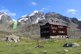 Geschindelte Hütte mit grünweißen Fensterläden und grüner Fahne vor schneebedeckten Gipfeln.