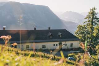 Alpenhütte mit dunklem Dach inmitten grüner Wiese und Bäumen, im Hintergrund hohe Berge und sanftes Sonnenlicht.
