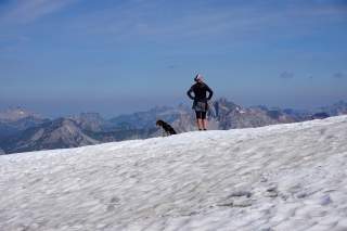 Wanderin und Hund stehen auf Schneefeld und blicken in die Ferne