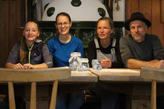 Eine Hüttenwirtsfamilie sitzt vor einem Kachelofen nebeneinander am Tisch.