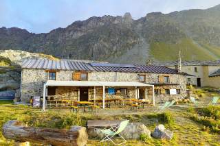 Hütte aus Steinmauern vor schroffer Bergkulisse, Sitzbänke und Liegestühle auf der Terrasse.