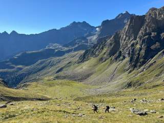 Zwei Wanderer steigen eine grüne, steinige Bergwiese hinauf, im Hintergrund hohe, felsige Berge unter blauem Himmel.