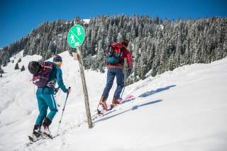 Zwei Skitourengeher steigen über eine verschneite Schneefläche bergauf an einem grünen Schild vorbei..