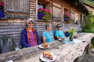 Drei Frauen sitzen mit Kuchen vor einer holzvertäfelten Almhütte