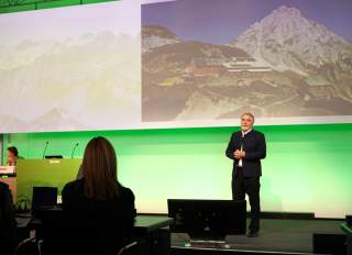 Ein Mann hält einen Vortrag vor einer Gruppe von Menschen, während im Hintergrund eine Projektion eines Bergpanoramas mit Hütten zu sehen ist.