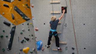 Ein Kind mit Klettergurt erklimmt eine Kletterwand und berührt oben ein markiertes Ziel.