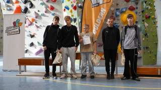 Fünf Jugendliche stehen in einer Kletterhalle vor einer Kletterwand und halten Taschen, einer hält eine Urkunde; im Hintergrund Banner der Deutschen Schulmeisterschaft im Klettern.