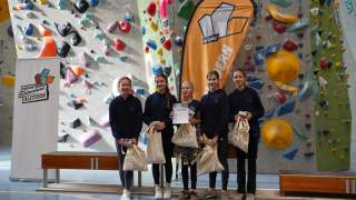 Fünf Mädchen stehen lächelnd mit Urkunden und Stofftaschen vor einer Kletterwand und Werbebannern bei einer Schulmeisterschaft im Klettern.
