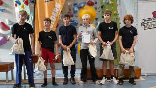 Sechs Jugendliche stehen vor einer Kletterwand, halten Stoffbeutel und eine Urkunde, einige tragen T-Shirts mit der Aufschrift "TJG".
