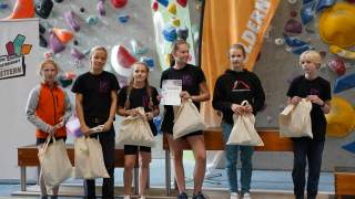Sechs Jugendliche stehen mit Stofftaschen in einer Kletterhalle vor einer Kletterwand, eine Person hält eine Urkunde, im Hintergrund Banner und bunte Klettergriffe.