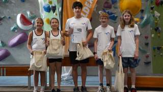 Fünf Kinder stehen in Sportkleidung vor einer Kletterwand und halten Stofftaschen, eines hält zudem eine Urkunde.