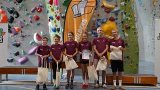 Sechs Jugendliche in einheitlichen bordeauxfarbenen T-Shirts stehen mit Stoffbeuteln und einer Urkunde vor einer Kletterwand und einem orangefarbenen Banner.