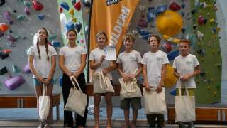Sechs Kinder und Jugendliche in weißen T-Shirts stehen mit Stofftaschen vor einer Kletterwand, eine Person hält eine Urkunde, im Hintergrund ein orangefarbener Banner mit der Aufschrift "Bouldern".
