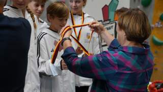 Eine Frau legt einem Jungen bei einer Siegerehrung eine Medaille mit schwarz-rot-gelbem Band um, weitere Jugendliche in weißen Sportjacken stehen daneben.