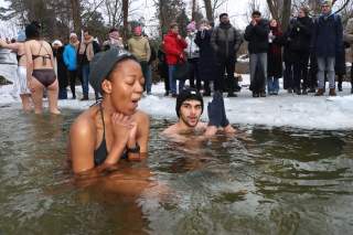 Zwei Menschen in Badekleidung sitzen im eisigen Wasser eines zugefrorenen Sees, während eine Gruppe warm angezogener Zuschauer am Ufer steht.