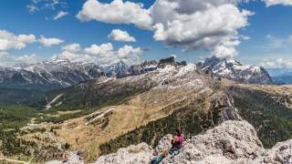 Frau sitzt auf einem Felsen und blickt auf eine weite Berglandschaft mit schneebedeckten Gipfeln und grünen Tälern unter blauem Himmel mit Wolken.