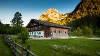 Ein altes Holzhaus mit einem Holzzaun, im Hintergrund Berge in der Abendsonne