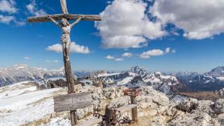Großes Holzkreuz mit Christusfigur auf schneebedecktem Berggipfel, im Hintergrund Panorama der Alpen unter blauem Himmel mit Wolken.