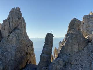 Eine Person steht auf einer schmalen Felsnadel zwischen steilen Felswänden in einer Gebirgslandschaft bei klarem Himmel.