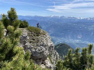 Ein Mountainbiker steht auf einem felsigen Bergvorsprung mit weitem Blick über ein Tal und schneebedeckte Berge im Hintergrund, umgeben von Kiefern.