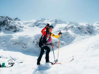 Skibergsteigerin mit Helm und bunter Kleidung erklimmt bei sonnigem Wetter und klarem Himmel eine schneebedeckte Berglandschaft.