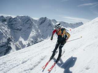 Skimo Athlet fährt den Berg runter. Es ist viel Schnee zu sehen, im Hintergrund schneebedeckte Berge und blauer Himmel. Er trägt einen Rennanzug und einen schwarz weißen Helm.