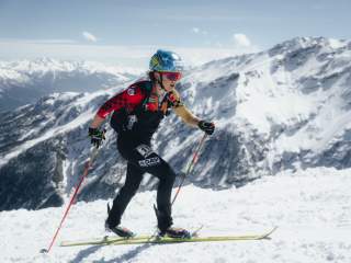 Skimo Athletin beim Aufstieg, im Hintergrund schneebedeckte Berge und blauer Himmel. Sie trägt einen Rennanzug, blauen Helm und neon pinke Sonnenbrille