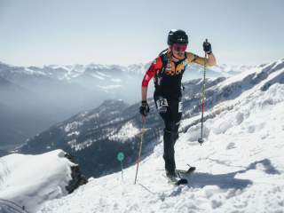 Skimo Athlet im Aufstieg. Im Hintergrund schneebedeckte und bewaldete Berge. Man sieht Sonnenstrahlen un
