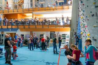In einer Kletterhalle mit blauem Boden sind viele Menschen, auch im Rollstuhl, um zu klettern.