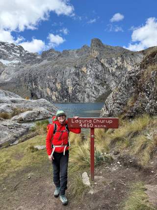 Frau mit roter Jacke steht neben einem Schild mit der Aufschrift „Laguna Churup 4.450 m.s.n.m.“ vor einem Bergsee und Felsen unter blauem Himmel.