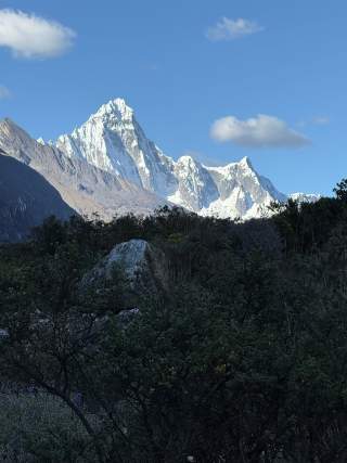Schneebedeckte Bergspitzen unter blauem Himmel mit vereinzelt weißen Wolken, im Vordergrund dichter grüner Wald und ein großer Felsen.