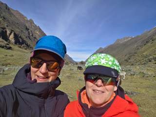 Zwei Frauen mit Outdoor-Bekleidung und Sonnenbrillen machen ein Selfie in einem grünen, bergigen Tal unter blauem Himmel. Im Hintergrund sind einige Kühe oder Rinder auf der Wiese zu sehen.