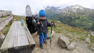 Kleinkind mit Wanderstöcken in der Hand, im Hintergrund die Berge und die Kraxe.