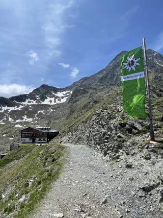 Wanderweg in den Bergen mit einer Alpengasthütte links und einer grünen Fahne mit Edelweißlogo rechts, umgeben von felsigem Gelände und schneebedeckten Gipfeln unter blauem Himmel.