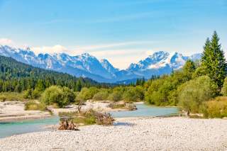 Blick auf eine breite Schotterfläche, in der ein Fluss mäandert. Im Hintergrund die Alpengipfel von Karwendel und Wetterstein.