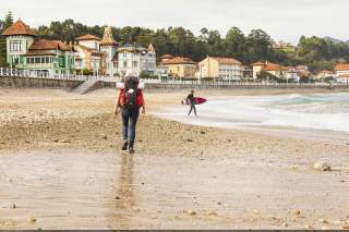 Wanderin läuft über einen Kiesstrand, ein Surfer geht mit seinem Board ins Meer.