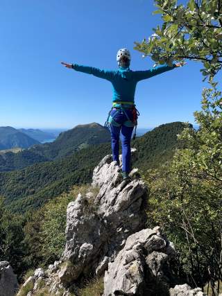 Eine Person mit Kletterausrüstung und Helm steht auf einem Felsen, breitet die Arme aus und blickt auf bewaldete Berge unter blauem Himmel.