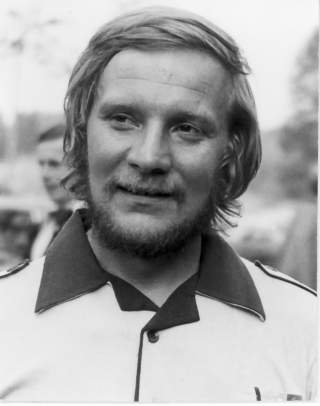 Porträt eines lächelnden Mannes mit Bart und mittellangem Haar, der ein Hemd mit dunkel abgesetztem Kragen trägt.