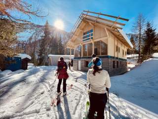 Drei Menschen mit Ski stehen im Schnee vor der Jugendherberge Fragant. Die Hütte aus Holz mit Steinfundament ist noch leer. Auf dem Dach liegt Schnee. Die Sonne scheint vom wolkenlosen Himmel.