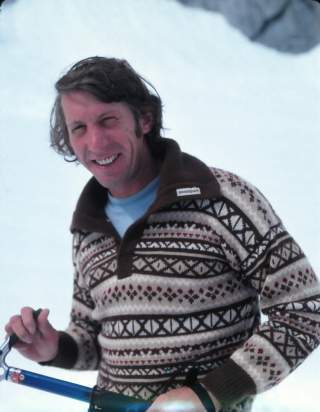 Mann mit lockigem Haar trägt einen gemusterten Pullover und hält einen Eispickel in verschneiter Landschaft.