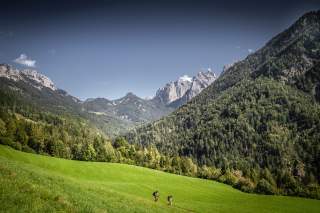 Zwei Wanderer mit Rucksäcken gehen auf einem grünen, hügeligen Feld, umgeben von bewaldeten Bergen unter einem klaren blauen Himmel.